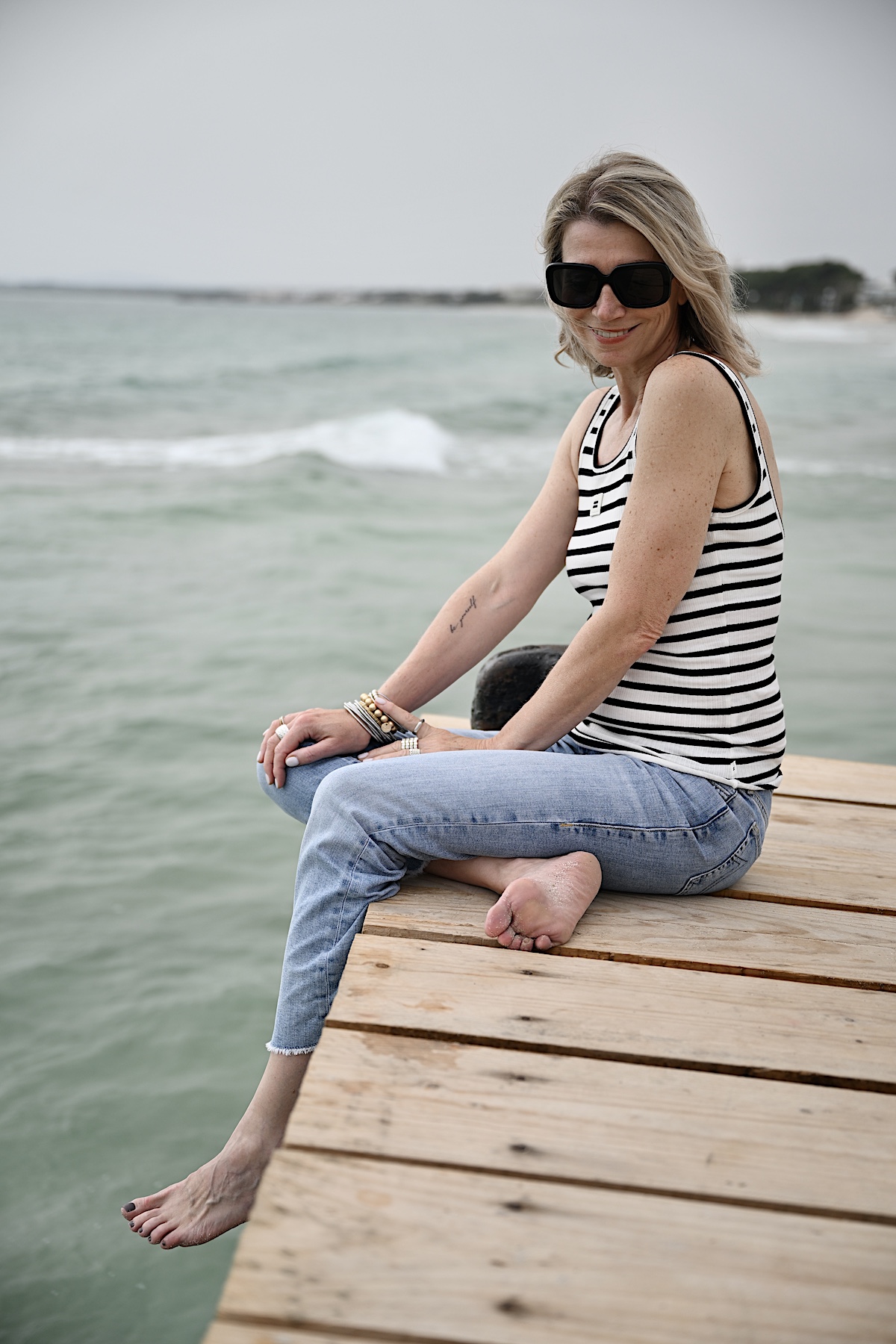 Beyond50. Sigi trägt eine helle destroyed Jeans , ein Streifentop mit Rückenausschnitt und eine schwarze Sonnenbrille, sitzt auf einem Steg am Meer.