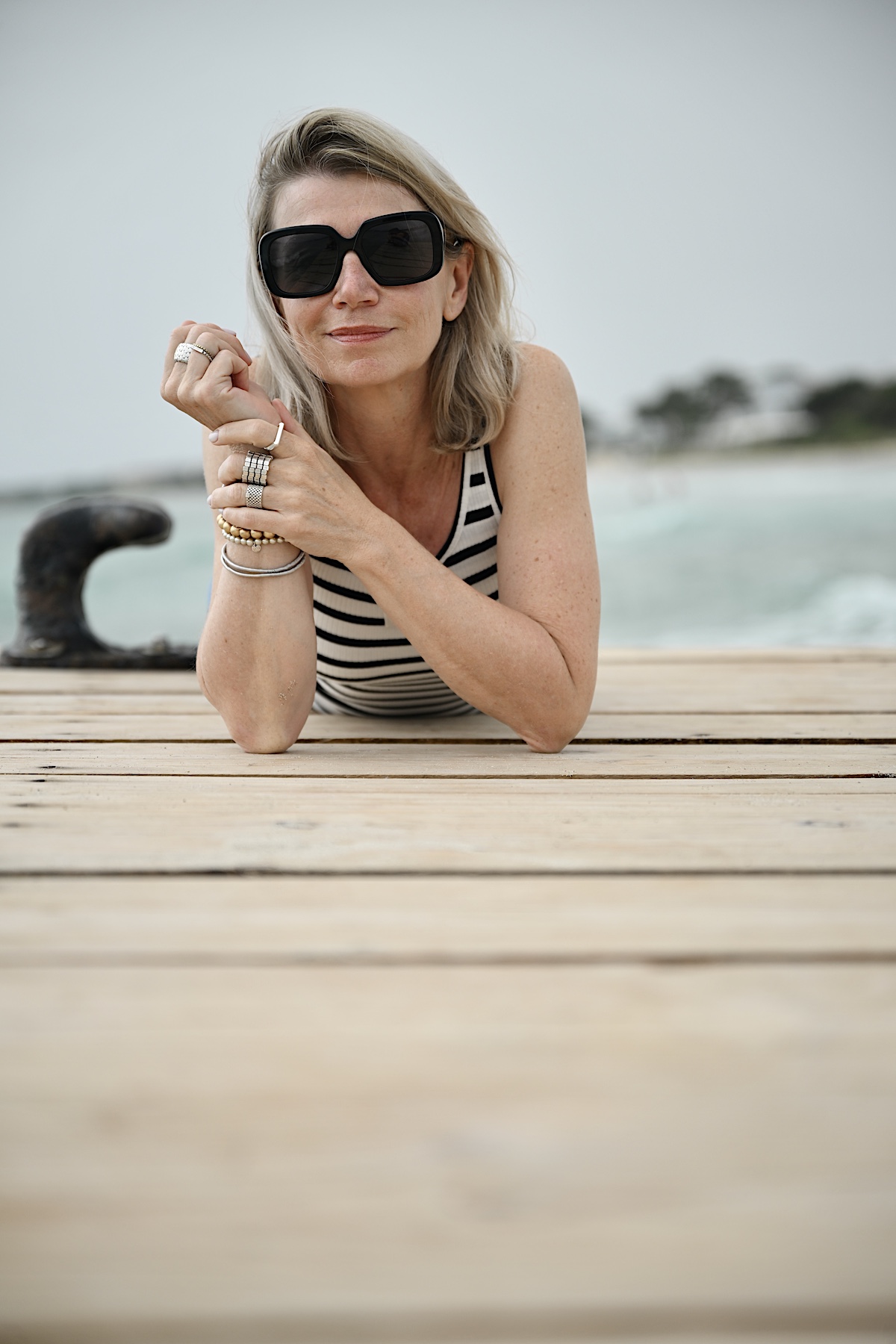 Beyond50. Sigi trägt eine helle destroyed Jeans , ein Streifentop mit Rückenausschnitt und eine schwarze Sonnenbrille, liegt bäuchlings auf einem Steg am Meer.