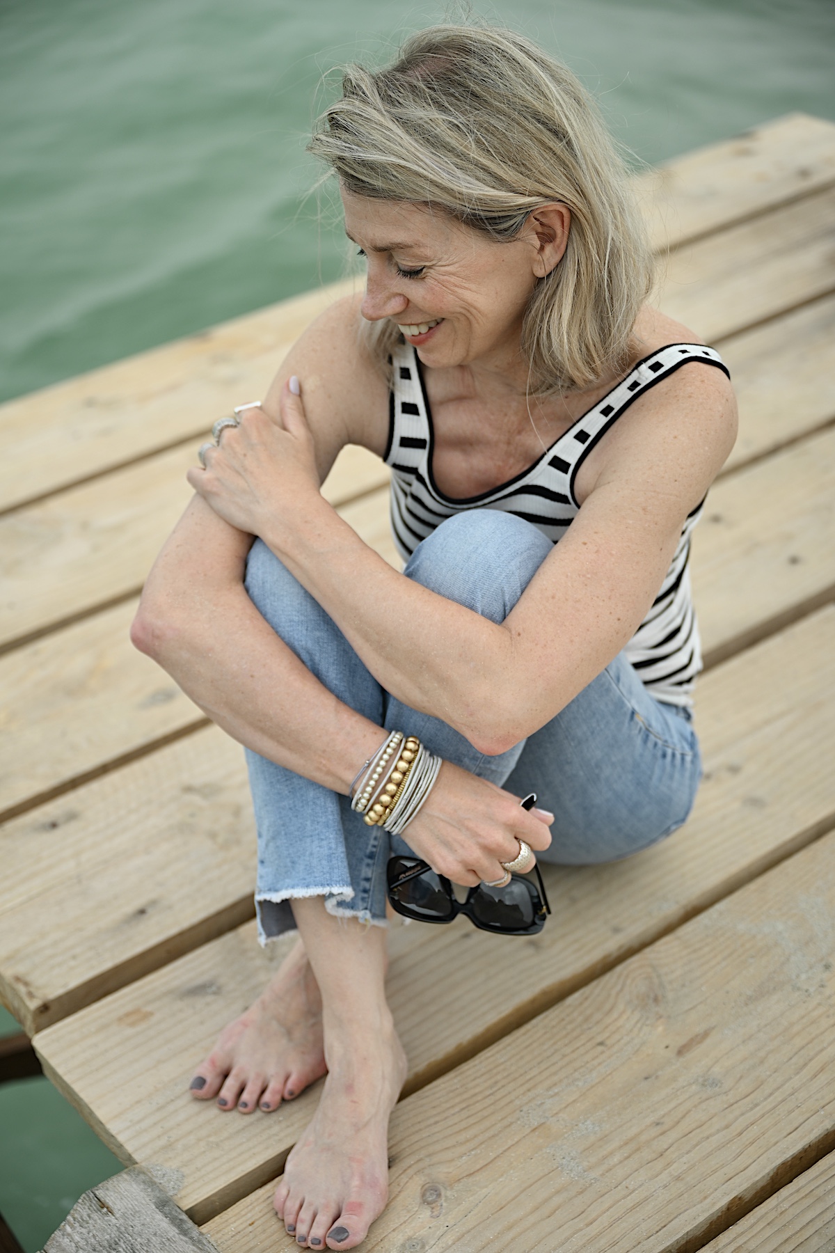 Beyond50. Sigi trägt eine helle destroyed Jeans , ein Streifentop mit Rückenausschnitt und eine schwarze Sonnenbrille, sitzt auf einem Steg am Meer. Knie von den Armen umschlungen.