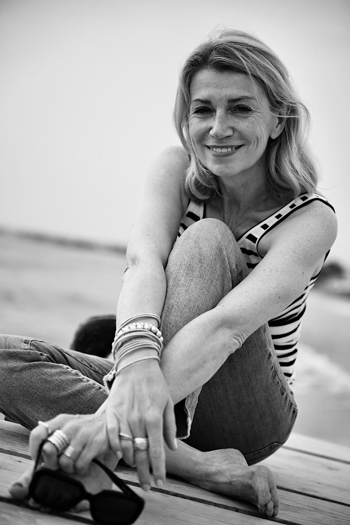 Beyond50. Sigi trägt eine helle destroyed Jeans , ein Streifentop mit Rückenausschnitt und eine schwarze Sonnenbrille in der Hand, sitzt auf einem Steg am Meer. SW.