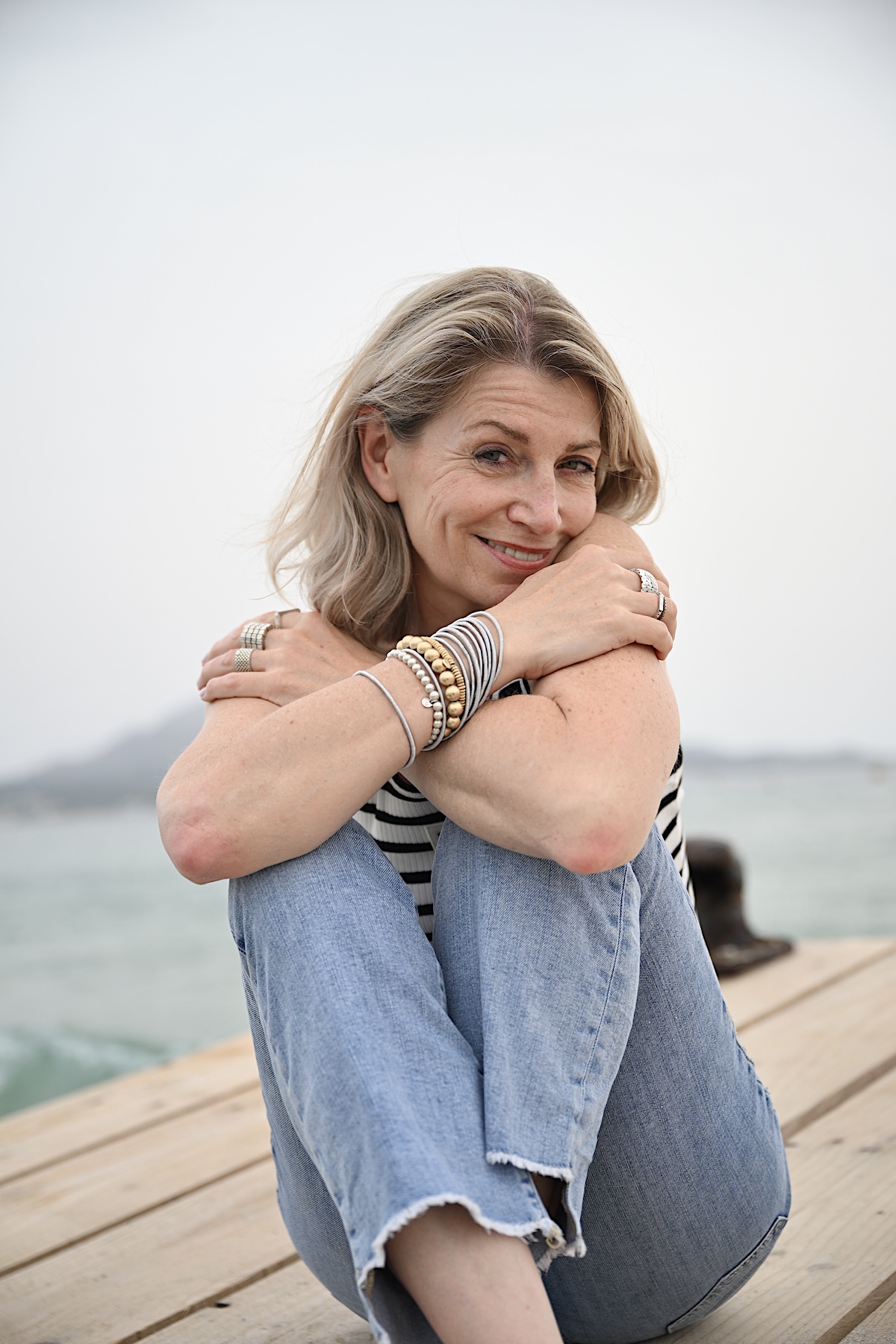 Beyond50. Sigi trägt eine helle destroyed Jeans , ein Streifentop mit Rückenausschnitt , sitzt auf einem Steg am Meer.