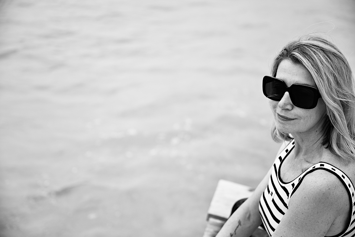 Beyond50. Sigi trägt eine helle destroyed Jeans , ein Streifentop mit Rückenausschnitt und eine schwarze Sonnenbrille, sitzt auf einem Steg am Meer. Portrait Ausschnitt.