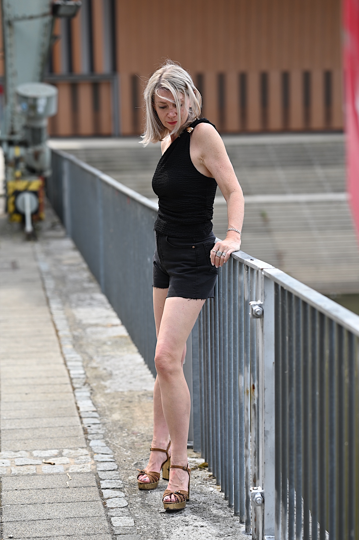 MindsetMondayShorts_KW33_1 Sigrid trägt schwarze Jeansshorts und dazu ein asymmetrisches Top auch schwarz mit einem goldenen Akzent, ähnlich einer Brosche, an dem einen Träger, dazu braune Wildledersandalen mit goldenem Plateau und Blockabsatz. Sie lehnt am Geländer am Main in Würzburg.