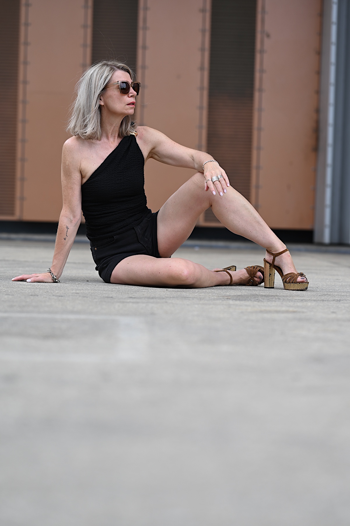 MindsetMondayShorts_KW33_10 Sigrid trägt schwarze Jeansshorts und dazu ein asymmetrisches Top auch schwarz mit einem goldenen Akzent, ähnlich einer Brosche, an dem einen Träger, dazu braune Wildledersandalen mit goldenem Plateau und Blockabsatz. Sie sitzt vor einer rostfarbenen Metallwand am Main in Würzburg.