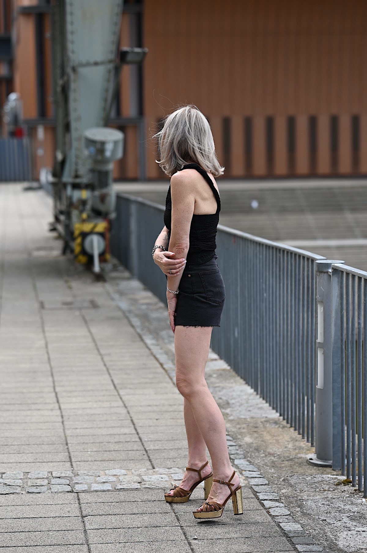 MindsetMondayShorts_KW33_2 Sigrid trägt schwarze Jeansshorts und dazu ein asymmetrisches Top auch schwarz mit einem goldenen Akzent, ähnlich einer Brosche, an dem einen Träger, dazu braune Wildledersandalen mit goldenem Plateau und Blockabsatz. Sie steht neben Geländer am Main in Würzburg.