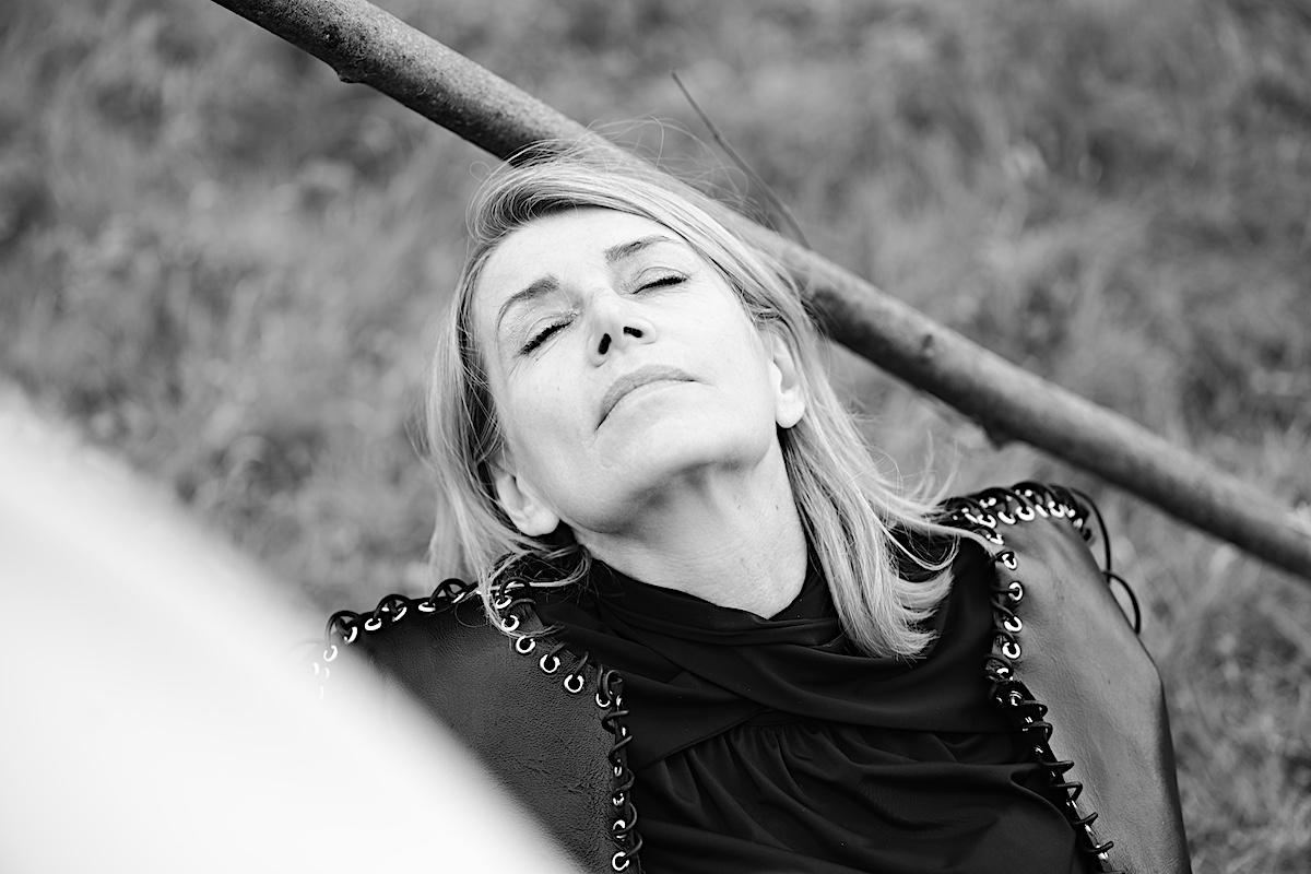 MindsetMonday Zeit. Portrait sw, outdoor, schwarze Bluse, Lederjacke mit Ösen und groben Lederband zusammengenäht. Sigrid sitzt am Boden, Kopf im Nacken. Augen geschlossen.