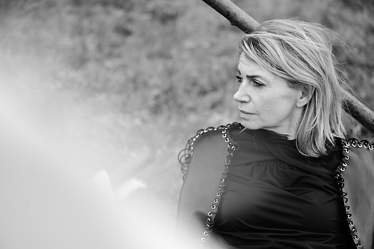 MindsetMonday Zeit. Portrait sw, outdoor, schwarze Bluse, Lederjacke mit Ösen und groben Lederband zusammengenäht. Sigrid sitzt am Boden, Schaut zur Seite in die Ferne