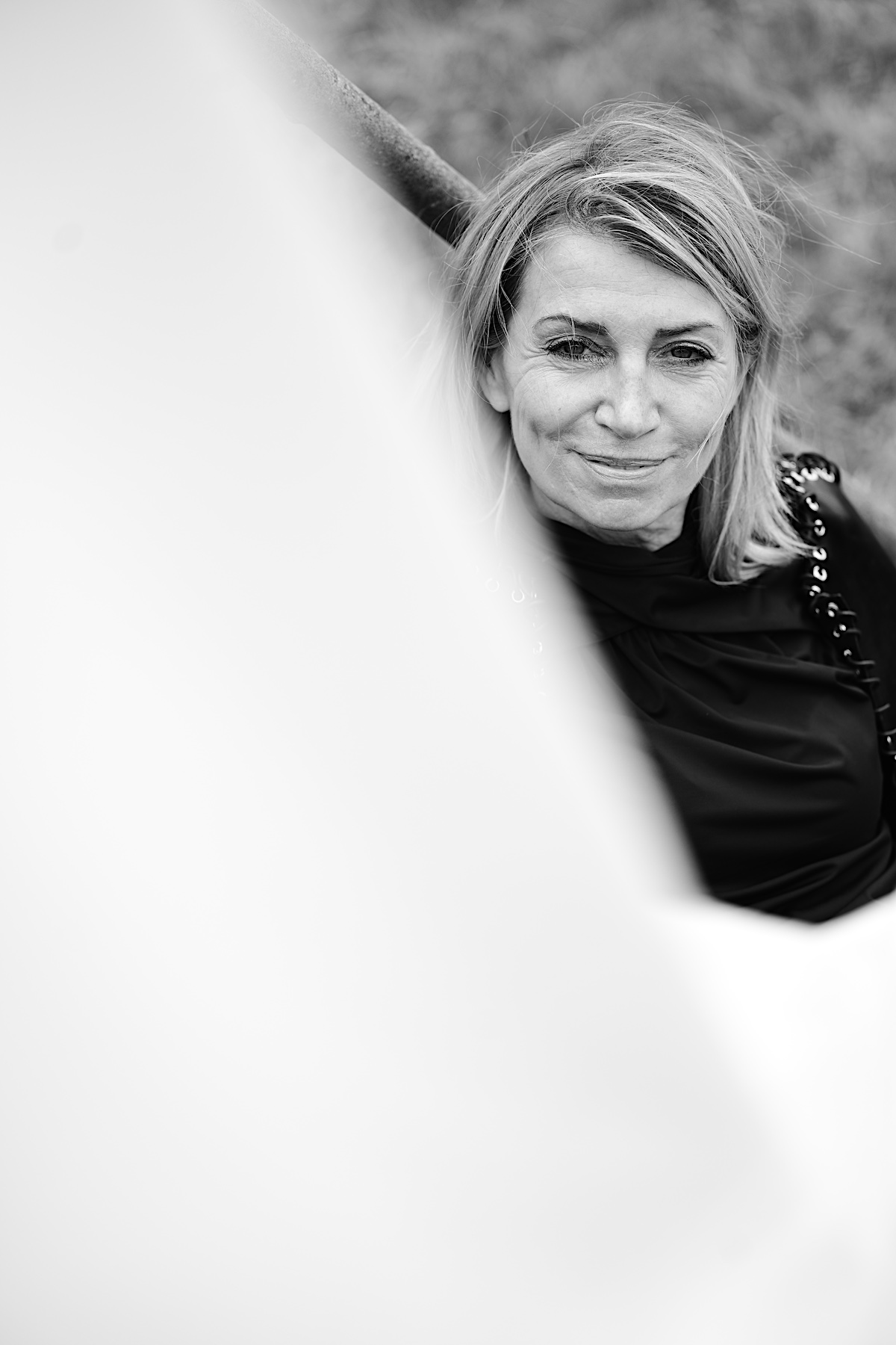 MindsetMonday Zeit. Portrait sw, outdoor, schwarze Bluse, Lederjacke mit Ösen und groben Lederband zusammengenäht. Sigrid sitzt am Boden, Schmunzelt mit direktem Blick in die Kamera