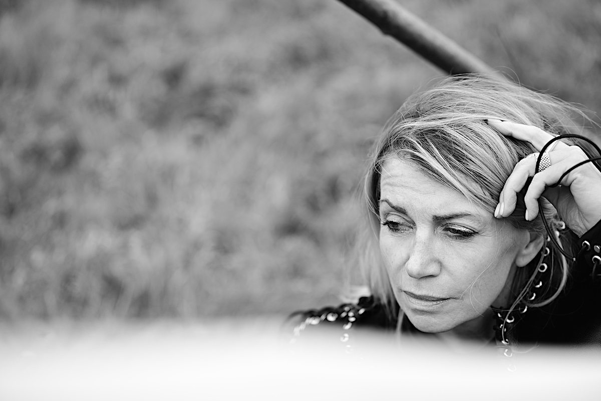 MindsetMonday Zeit. Portrait sw, outdoor, schwarze Bluse, Lederjacke mit Ösen und groben Lederband zusammengenäht. Sigrid sitzt am Boden, Hand in den Haaren, schaut ernst zur Seite