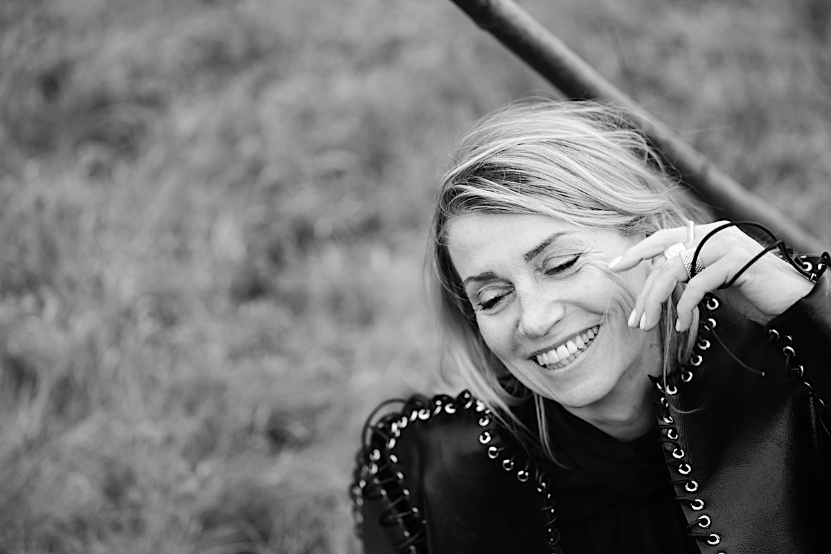MindsetMonday Zeit. Portrait sw, outdoor, schwarze Bluse, Lederjacke mit Ösen und groben Lederband zusammengenäht. Sigrid sitzt am Boden, Hand an der Wange, Augen geschlossen, lacht herzhaft