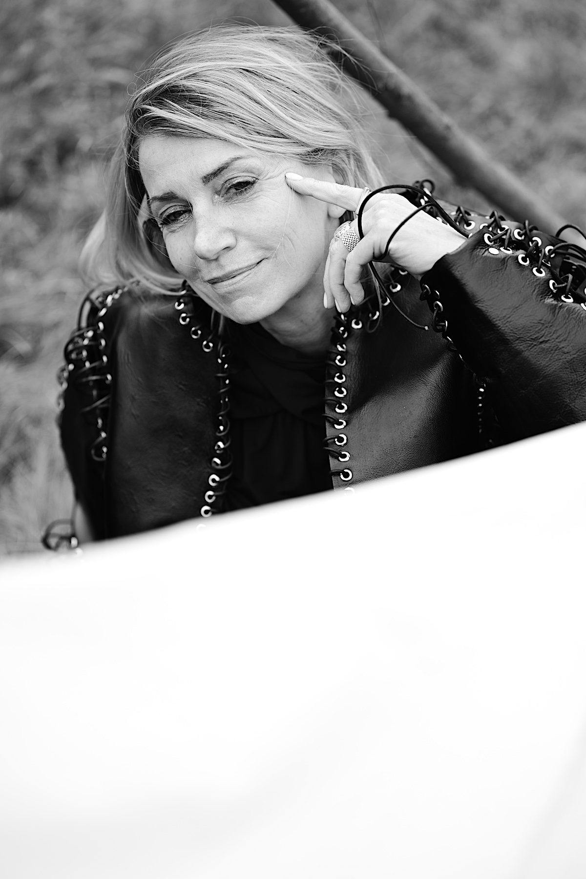 MindsetMonday Zeit. Portrait sw, outdoor, schwarze Bluse, Lederjacke mit Ösen und groben Lederband zusammengenäht. Sigrid sitzt am Boden, Hand an der Wange, lächelt in die Kamera