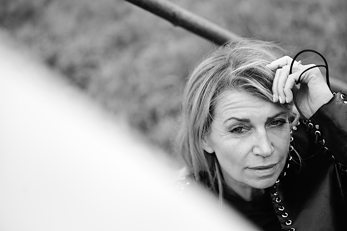MindsetMonday Zeit. Portrait sw, outdoor, schwarze Bluse, Lederjacke mit Ösen und groben Lederband zusammengenäht. Sigrid sitzt am Boden, Hand in den Haaren,nachdenkliche Pose